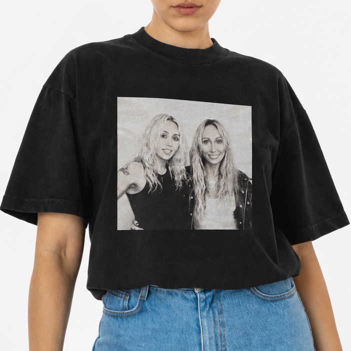 A woman modeling a black  Miley Cyrus and Tish Cyrus-Purcell graphic t-shirt with a black-and-white photo print on the front, paired with light blue denim shorts, against a plain white studio background.